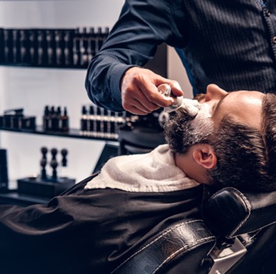 A man in a barbers chair having shaving cream applied with a brush above his beard, with a back drop of hair products on shelving