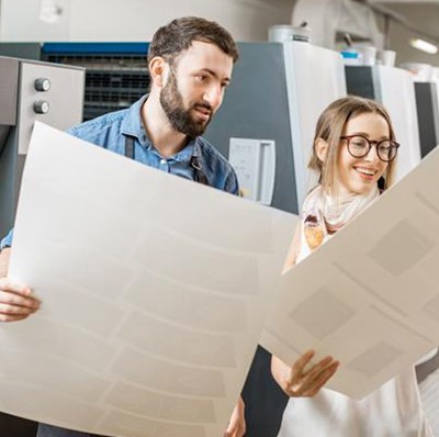 A man and a women each holding and looking A1 printed cards with  a row of industrial printers behind them A man and a women each holding and looking A1 printed cards with  a row of industrial printers behind them