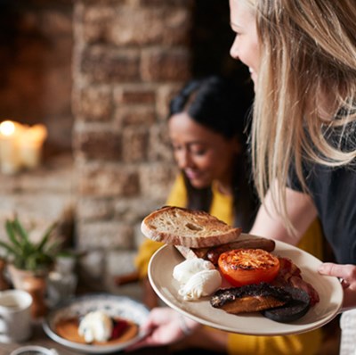 A waitress serving two fried breakfasts to a table with a large brick fireplace containing a wood fired stove in the background