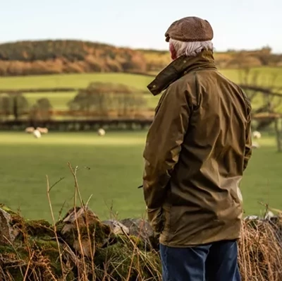 Man with grey hair and waxed jacket standing by a low dry wall with a blurred back drop of sheep in a field, trees and fields against a light blue sky Man with grey hair and waxed jacket standing by a low dry wall with a blurred back drop of sheep in a field, trees and fields against a light blue sky