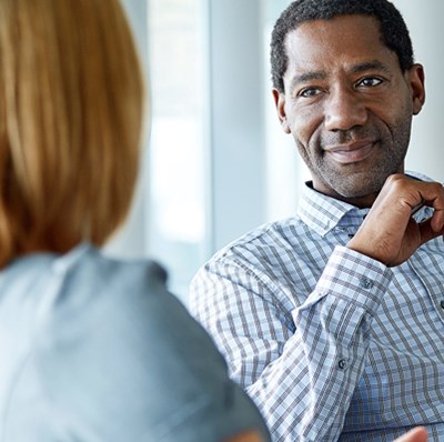 A head and shoulder view of a man in a checked shirt with a pen in his hand smiling at a women who we see the back of A head and shoulder view of a man in a checked shirt with a pen in his hand smiling at a women who we see the back of