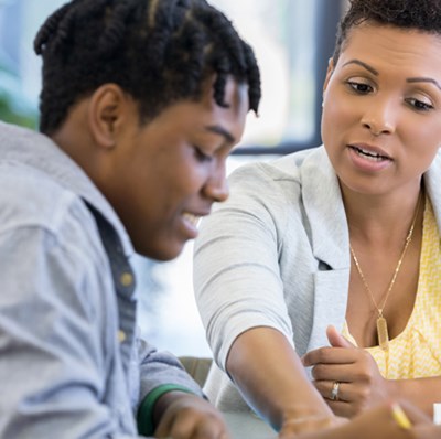 Head and shoulder view of a young man sitting beside a women while they work on something he is writing on at a desk with a blurred plant in background Head and shoulder view of a young man sitting beside a women while they work on something he is writing on at a desk with a blurred plant in background