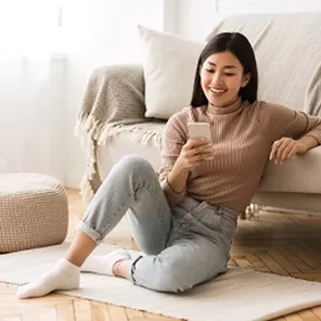 Smiling women sitting on a rug on a parquet floor leaning on a sofa looking at her mobile Smiling women sitting on a rug on a parquet floor leaning on a sofa looking at her mobile