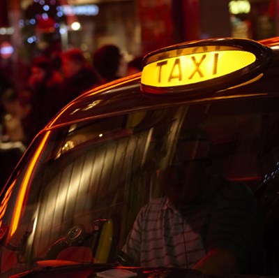 Lit taxi sign light on top of a modern London black cab at night, with a blurred background of some people and lights