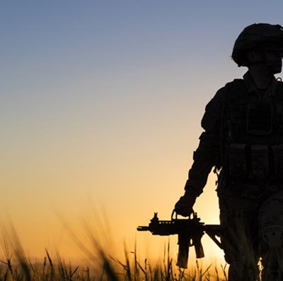 Silhouette of a soldier standing in grass holding a machine gun at his thigh with a sunset Silhouette of a soldier standing in grass holding a machine gun at his thigh with a sunset