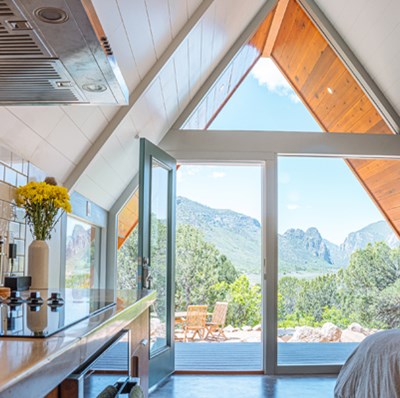 Bedsit with a peaked roof framed glass wall with an open door, a view of wooden patio furniture, mountains and greenery backdrop Bedsit with a peaked roof framed glass wall with an open door, a view of wooden patio furniture, mountains and greenery backdrop