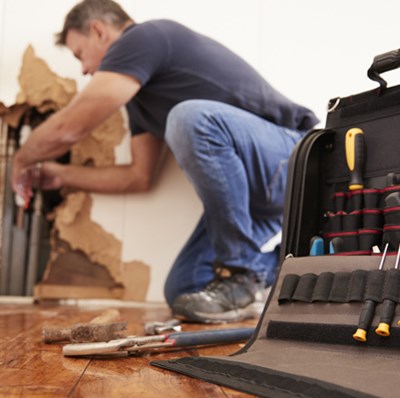 A man on one knee while he works on some exposed pipes in the corner of a room, his open tool bag in focus in the foreground on the right