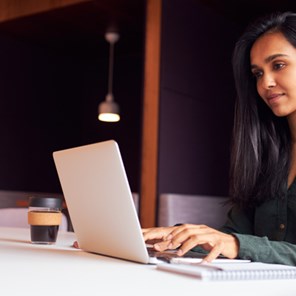 Woman typing on a laptop at a table with a coffee and notebook, a blurred backdrop of break out spaces with seats Woman typing on a laptop at a table with a coffee and notebook, a blurred backdrop of break out spaces with seats