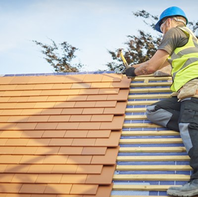 Man in a hard hat and high vis, lying sideways on the pitched roof of a new building, adding tiles with a hammer