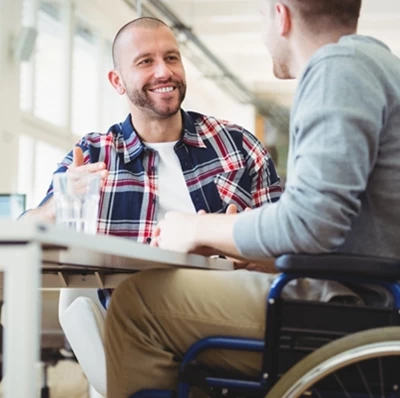 Man in a wheel chair at a desk talking to a man who is smiling sitting next to him