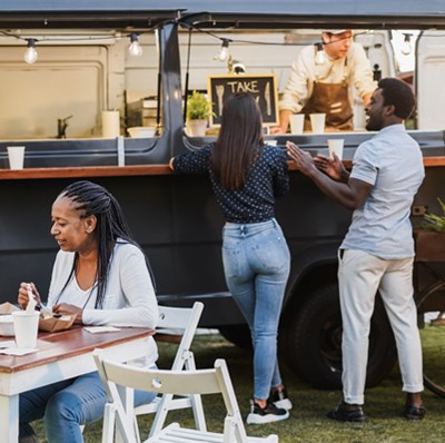 Three people queuing beside a mobile van with side hatches open serving take away food with two women enjoying their food at a nearby table Three people queuing beside a mobile van with side hatches open serving take away food with two women enjoying their food at a nearby table