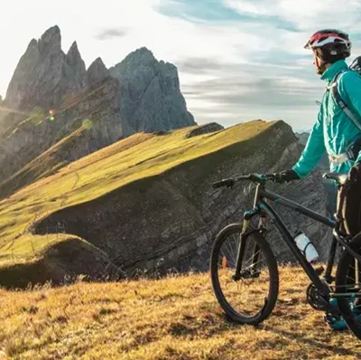 A man stood next to his bike on top of a grassy mountain looking at the rocky tops of other mountains  A man stood next to his bike on top of a grassy mountain looking at the rocky tops of other mountains