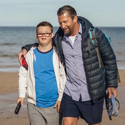 Man with an arm around the shoulders of a Downs syndrome boy with a labradoodle on a lead as they walk along a sandy beach