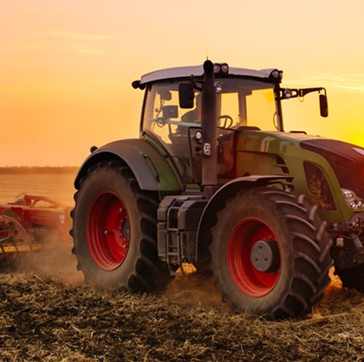 A tractor ploughing a field of soil with a backdrop of a low sun and yellow pinkish sky  