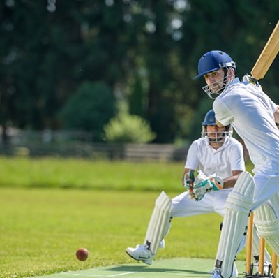 Batsman swinging his bat with the wicket keeper poised to catch the ball with a blurred tree lined backdrop Batsman swinging his bat with the wicket keeper poised to catch the ball with a blurred tree lined backdrop
