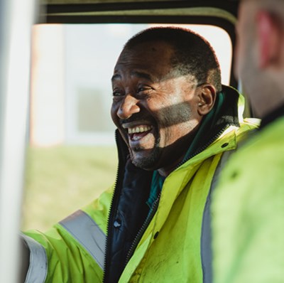 Side window view of a laughing man driving a van with the passenger facing towards him, both wearing yellow hi vis jackets
