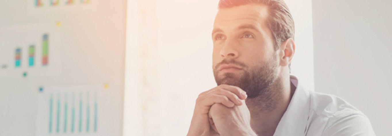 Man looking up thoughtfully, with clasped hands beside his chin and a back drop of graphs on a wall