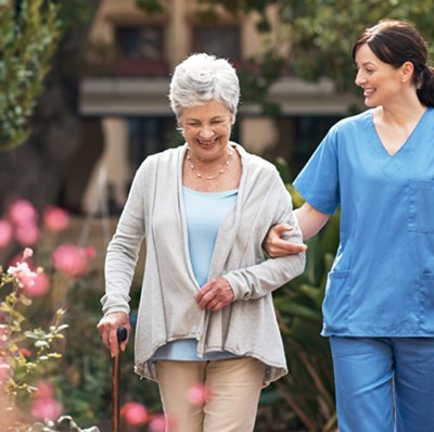 Older women walking with a stick in a garden of flowers and bushes with a lady in blue scrubs with her hand through the ladies arm