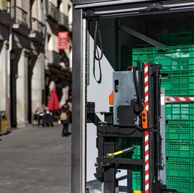 Green plastic food crates stacked in a van with a roller shutter door, a street view to the left hand side of stone buildings on a wide footpath Green plastic food crates stacked in a van with a roller shutter door, a street view to the left hand side of stone buildings on a wide footpath